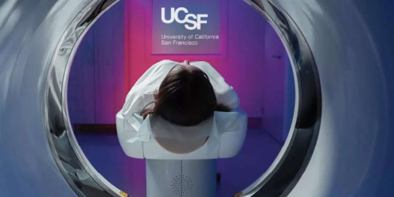 View from inside an MRI scanner looking outward at a patient lying on the table, facing a wall lit with purple and blue light and a UCSF logo projected in front of them.