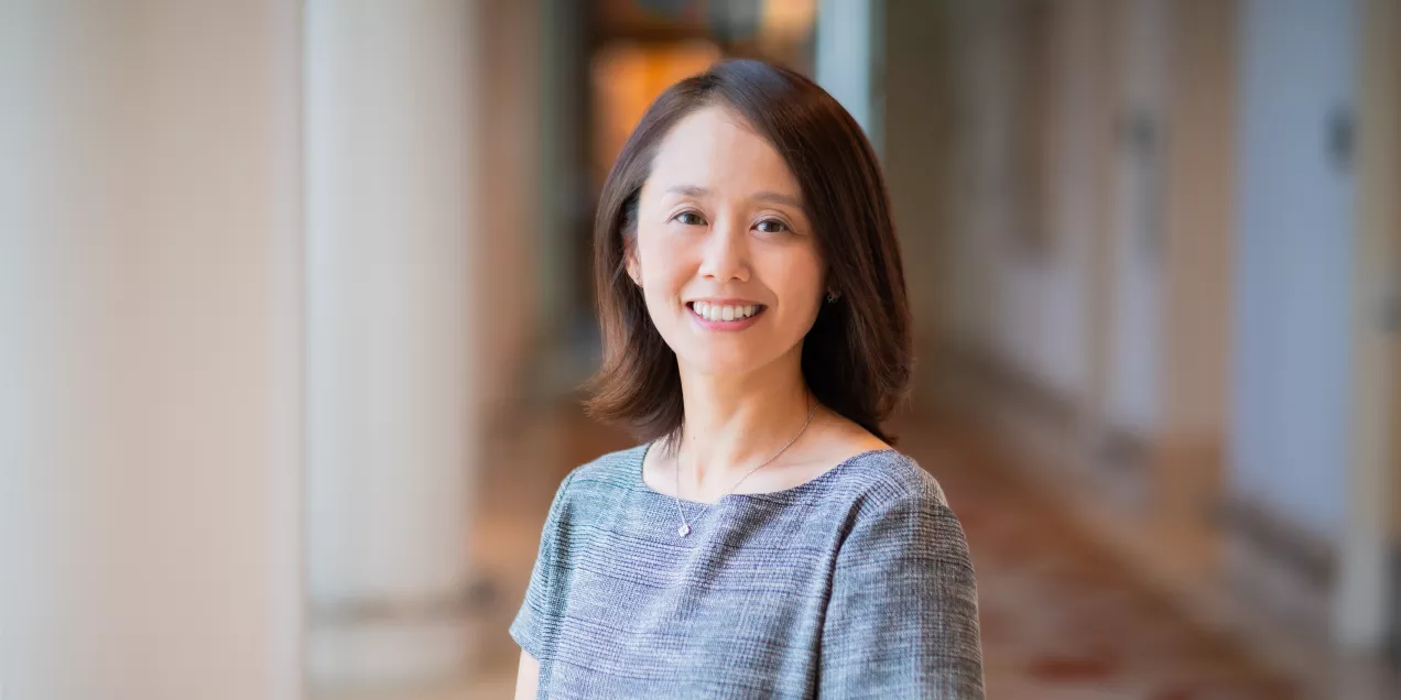 Natsuko Yamashita smiling while standing in a softly lit hallway with columns and patterned flooring, wearing a short-sleeved gray top.