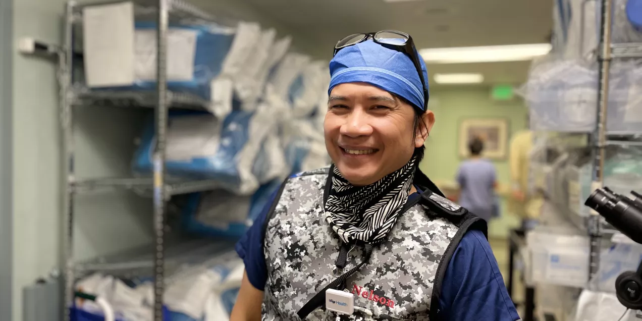A medical professional wearing a patterned protective vest, blue surgical cap, and face covering stands smiling in a supply room lined with shelves of packaged medical materials, with other staff visible in the background.