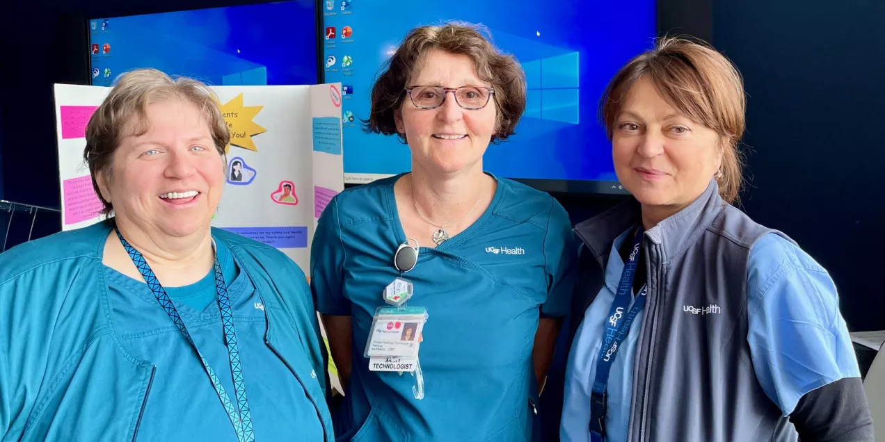 Three healthcare staff members standing together and smiling at an indoor event, with a display board and large monitors visible in the background.