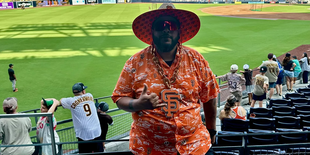 Rodolfo Baltodano standing in the stands at a baseball stadium wearing a bright orange San Francisco Giants shirt and wide-brimmed hat, posing with a shaka hand sign as the field and crowd sit behind him.