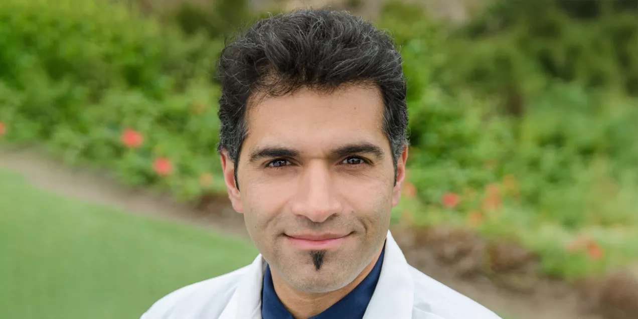 Sawhney Rajiv wearing a white lab coat, navy shirt, and blue tie, standing outdoors with a blurred background of greenery and flowers. He is looking at the camera with a slight smile.