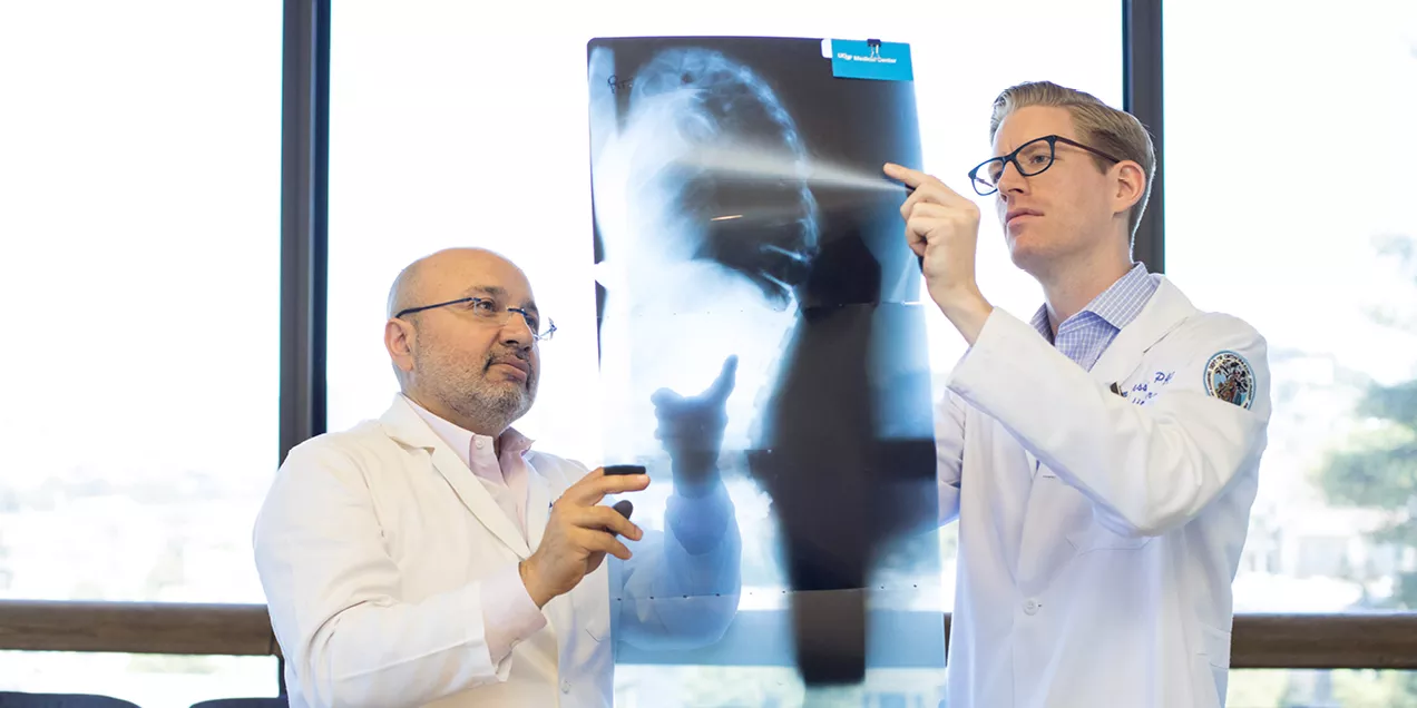 two doctors in white coats reviewing a large spinal X-ray held up to the light, discussing findings while standing in front of a bright window