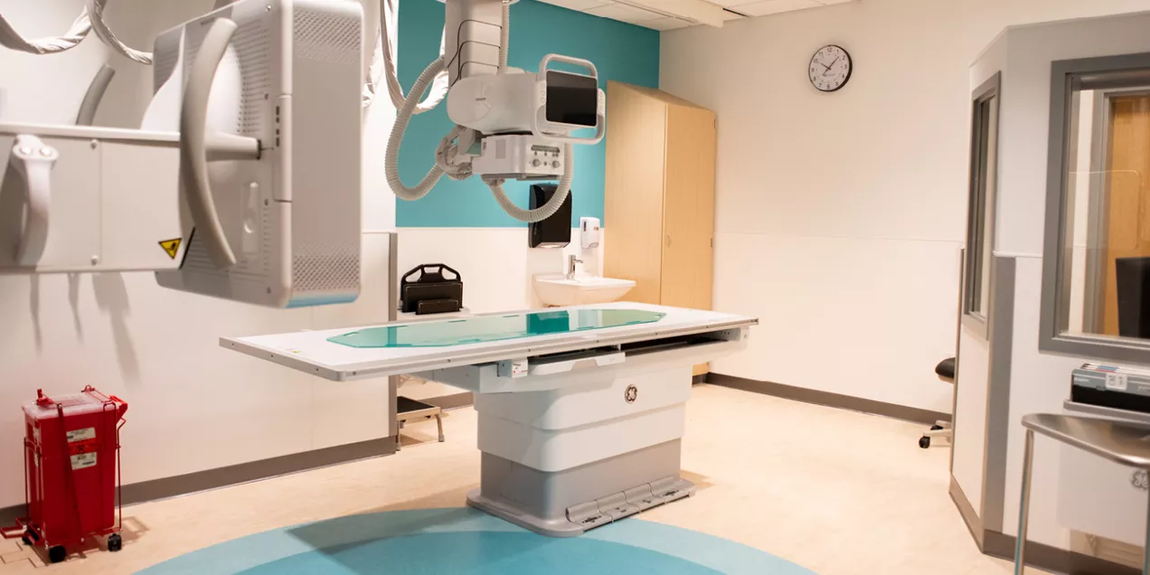 x-ray imaging room with a large radiography table, ceiling-mounted x-ray tube, detector panel, and surrounding medical equipment, shown in a clean clinical setting with teal and white walls