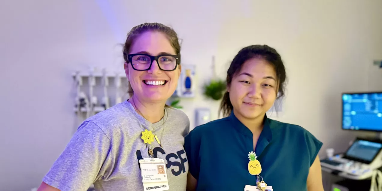 Two UCSF sonographers smiling together in a dimly lit exam room. One wears glasses and a UCSF t-shirt, and the other wears teal scrubs; both have ID badges with cheerful badge decorations. An ultrasound machine sits in the background.