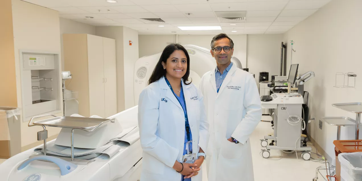 Two physicians in white lab coats standing in a hospital imaging room with a CT scanner and medical equipment in the background.