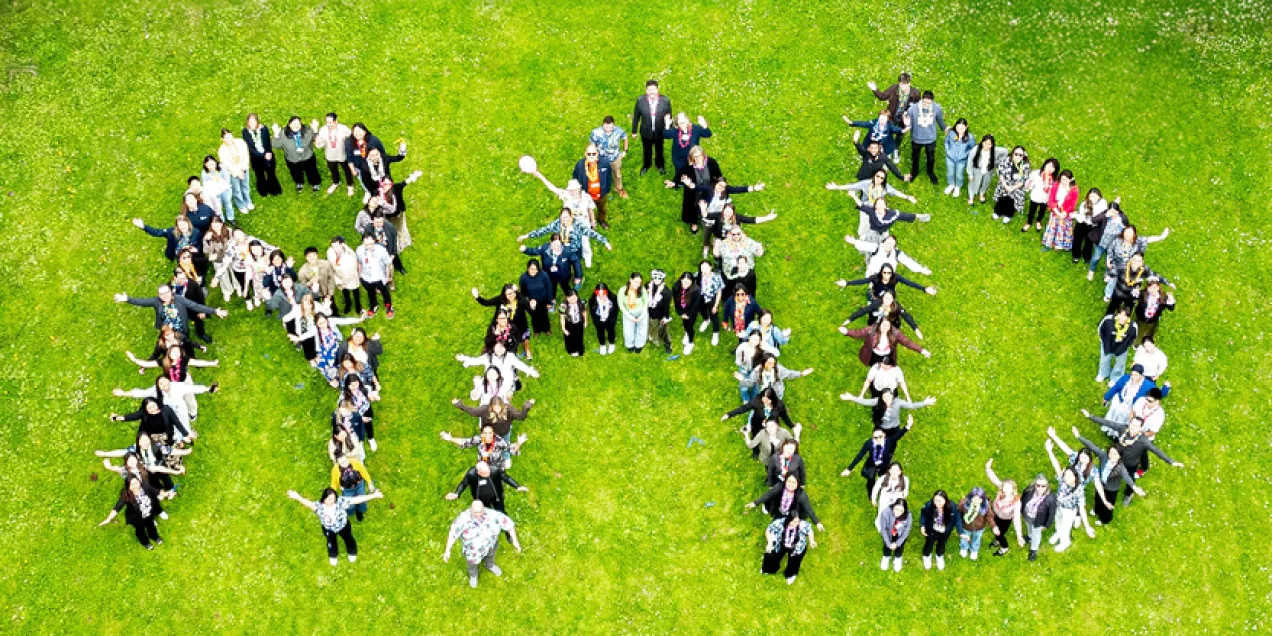 A large group of peopl e standing on a grassy field arranged to form the letters RAD, seen from above. The group is smiling and looking up with arms raised, creating a playful and energetic formation.