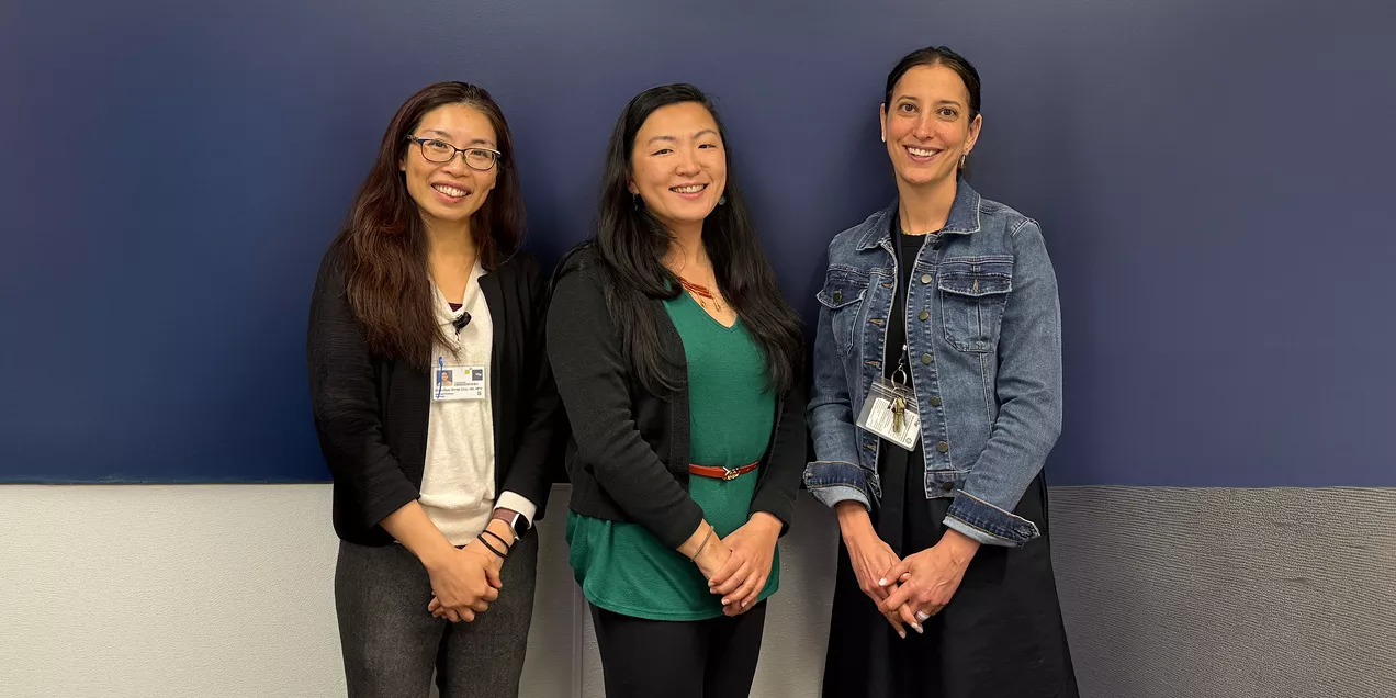 Three staff members stand side by side indoors against a blue and light gray wall, smiling at the camera, wearing business casual clothing with ID badges visible