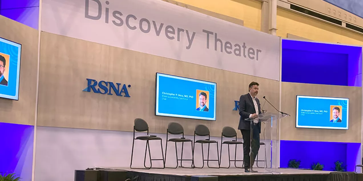 Christopher Hess stands at a podium speaking on a stage at the RSNA Discovery Theater, with large presentation screens displayed behind him.