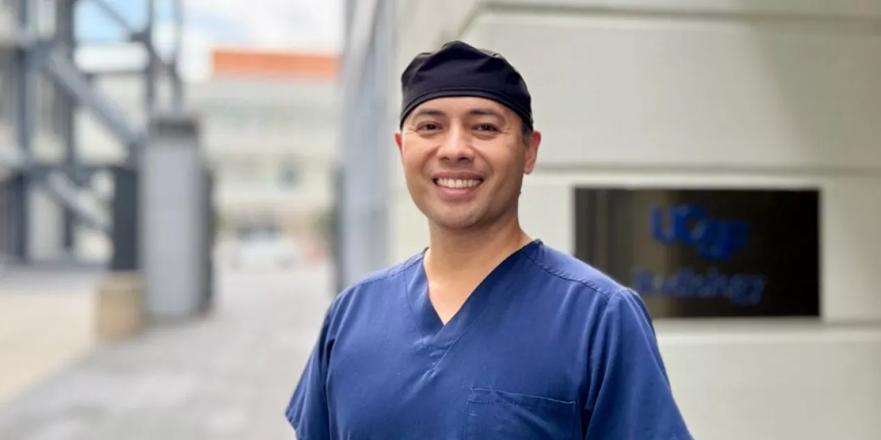 Portrait of a UCSF nurse wearing blue scrubs and a black surgical cap, smiling outdoors near a medical facility entrance.
