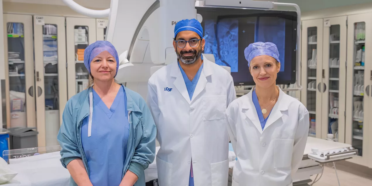 Three clinicians in surgical attire and white coats stand together in an interventional radiology suite, with medical equipment and imaging monitors visible behind them.