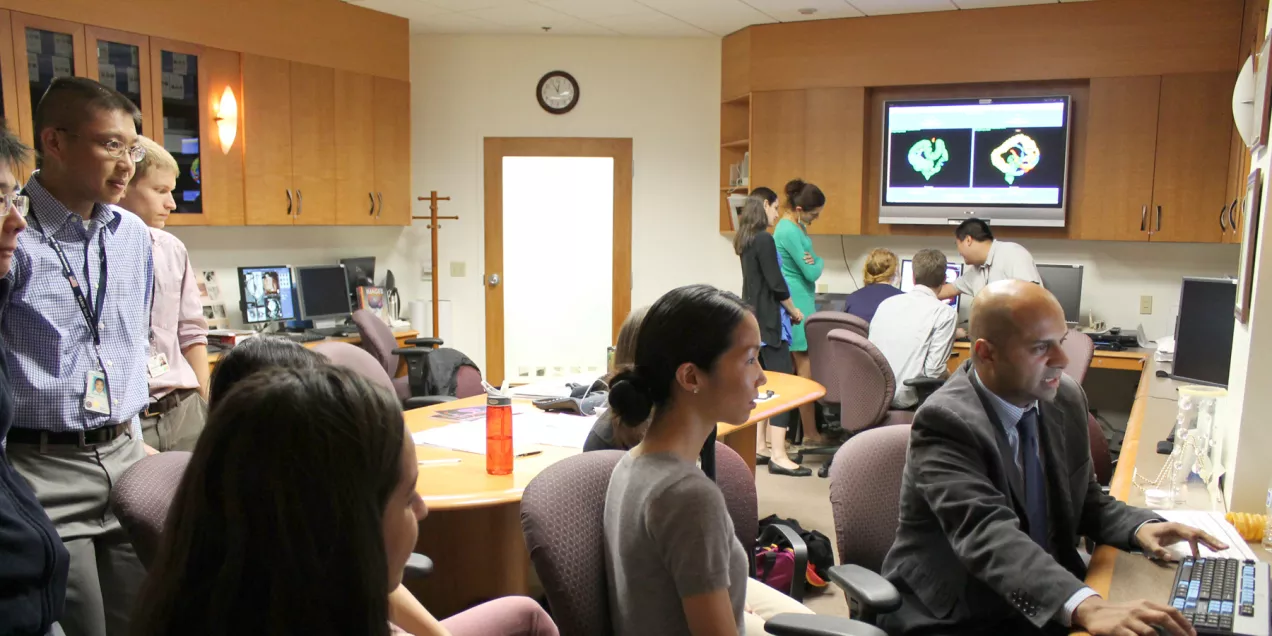 Medical trainees and clinicians gather in a radiology reading room, observing imaging studies displayed on computer monitors during a teaching session.