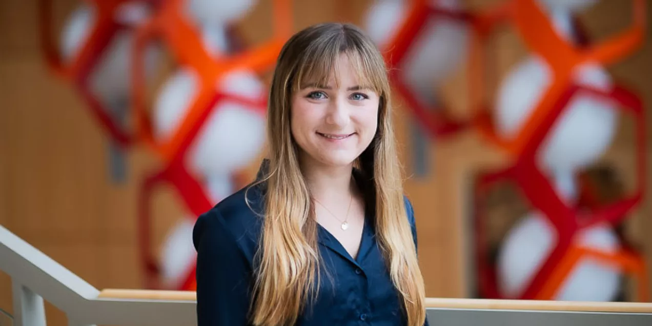 A woman with long blonde hair and bangs wearing a dark blouse stands indoors, smiling at the camera, with a softly blurred background featuring red geometric structures and warm tones.
