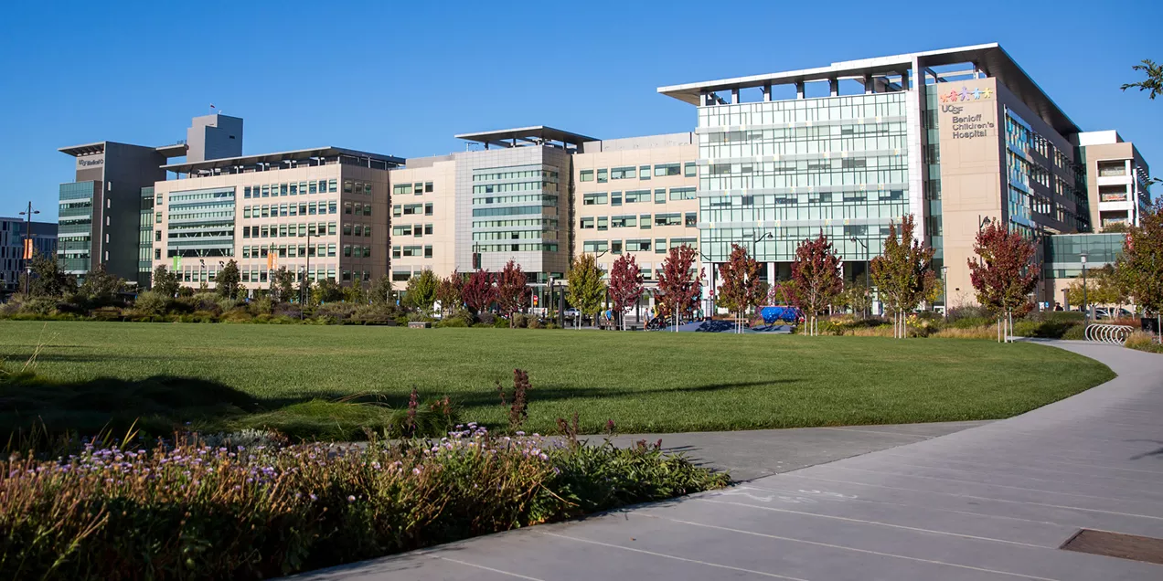 Wide exterior view of UCSF Benioff Children’s Hospital at Mission Bay, showing the full hospital complex with glass and stone facades, landscaped lawn and walking path in the foreground, and trees with red foliage under a clear blue sky.
