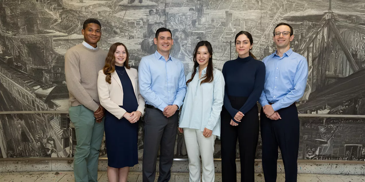 Five neuro-interventional fellows standing together in front of a large grayscale mural, including one physician in a white coat and blue scrubs, with the group facing the camera and smiling.