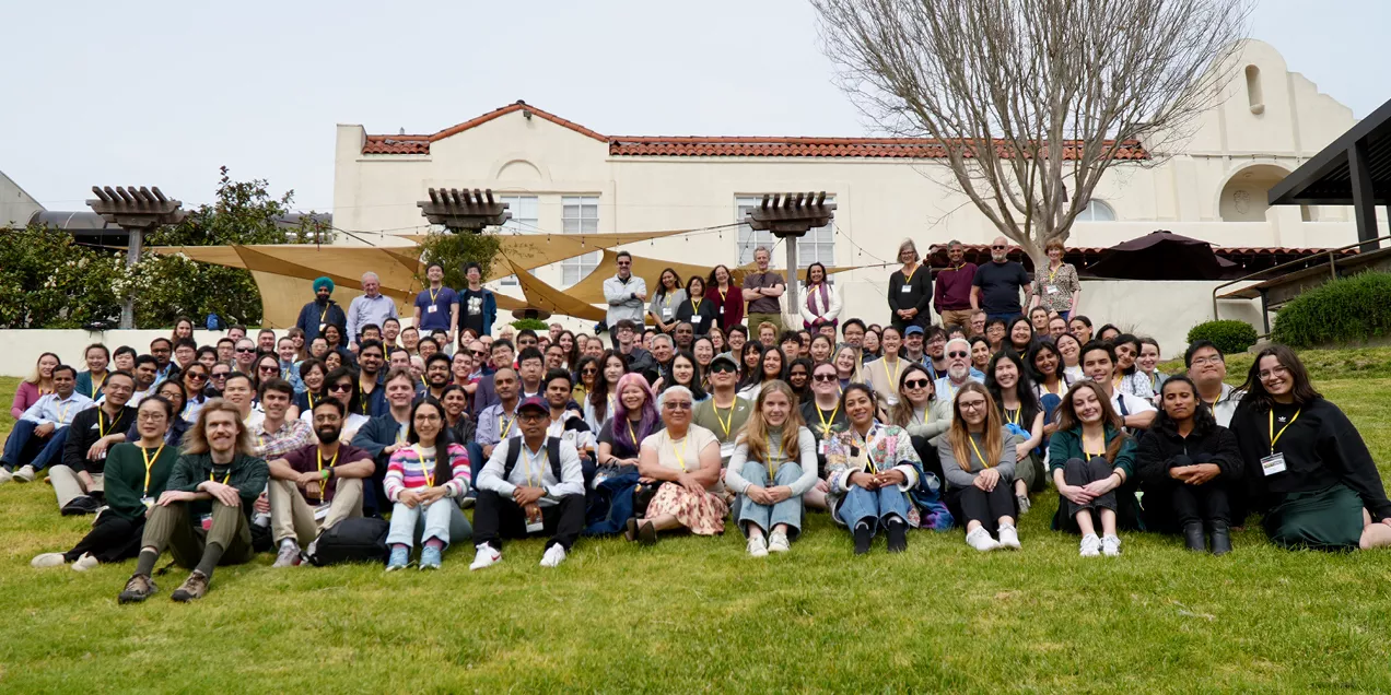 Large group photo of attendees seated and standing on a grassy lawn in front of a building during a research conference.