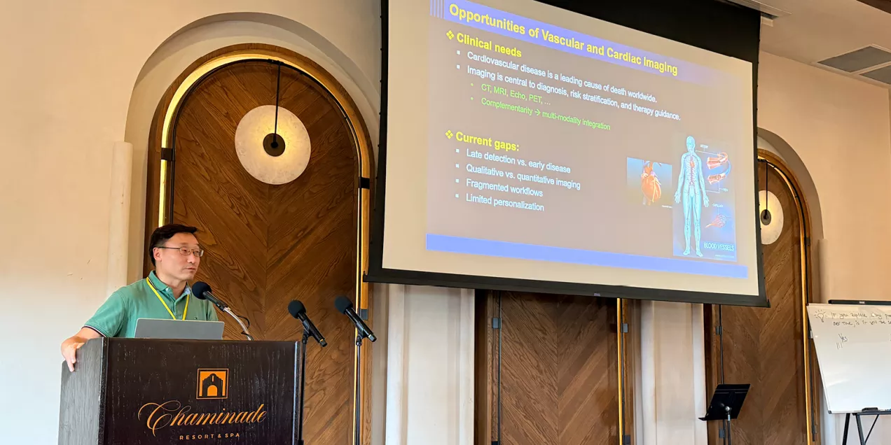 Speaker presenting at podium with projected slide on vascular and cardiac imaging research at conference