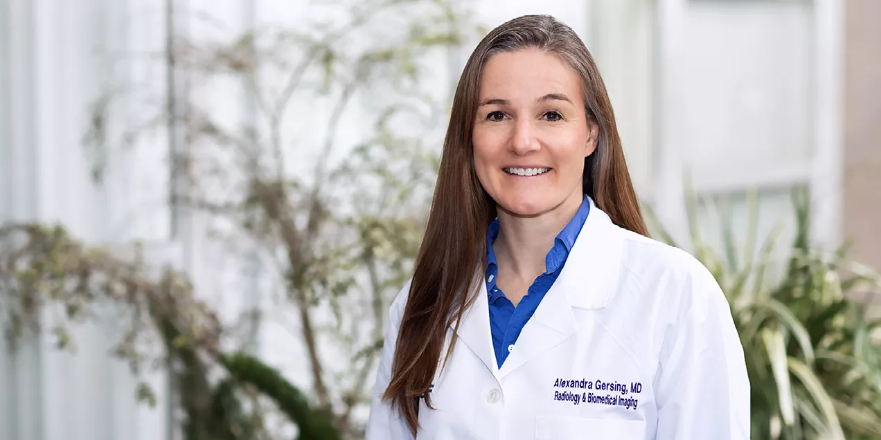 Portrait of a female physician in a white lab coat embroidered with “Alexandra Gersing, MD, Radiology & Biomedical Imaging,” smiling and standing indoors with soft natural light and greenery in the background.