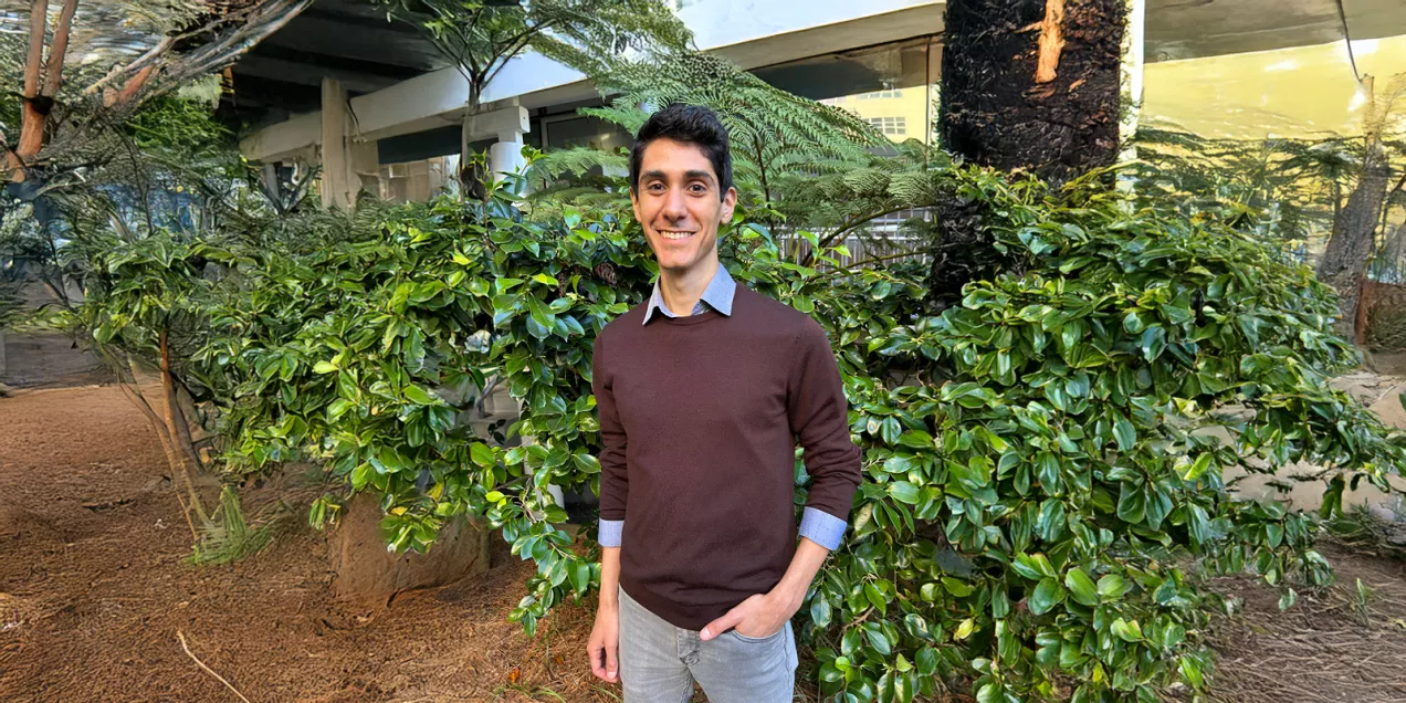 Portrait of a man smiling outdoors in a landscaped courtyard with greenery and trees, wearing a brown sweater and gray pants