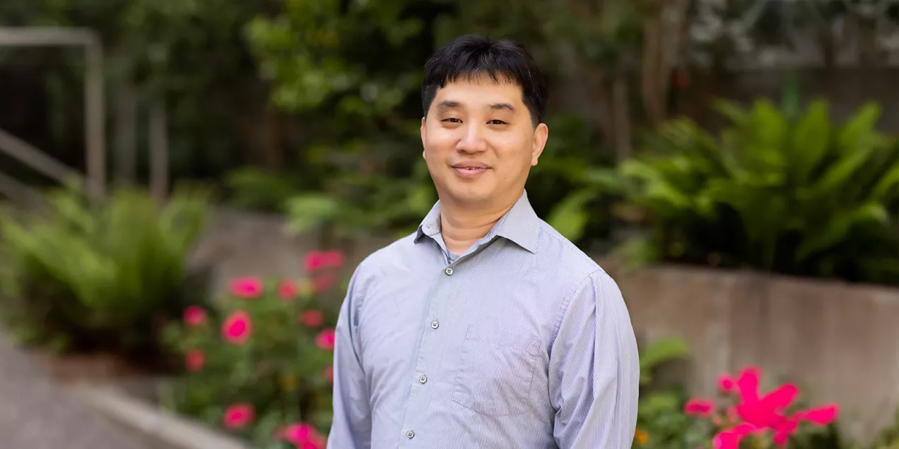 Portrait of a man with glasses smiling outdoors in a courtyard with greenery, wearing a black polo shirt