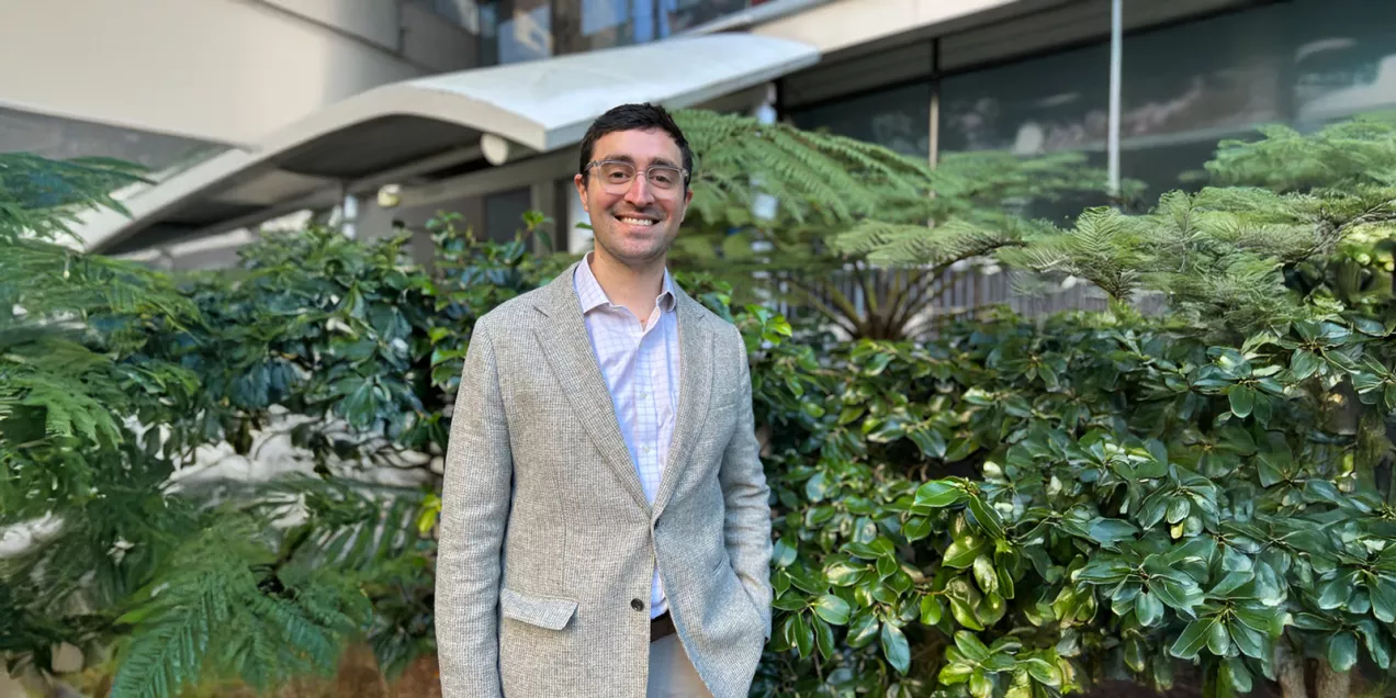 Portrait of a man smiling in an outdoor courtyard with lush plants, wearing glasses and a light gray blazer over a button-down shirt