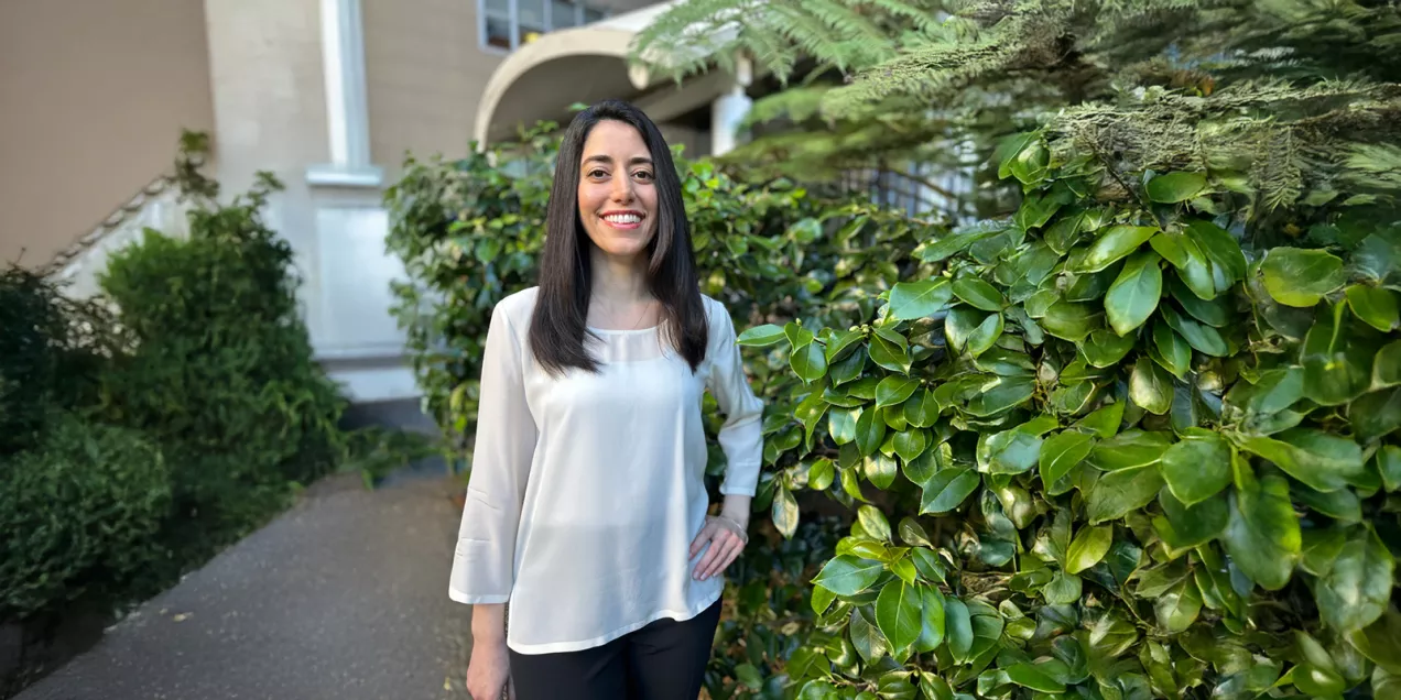 Portrait of a woman smiling in a garden walkway with greenery, wearing a light blouse and dark pants