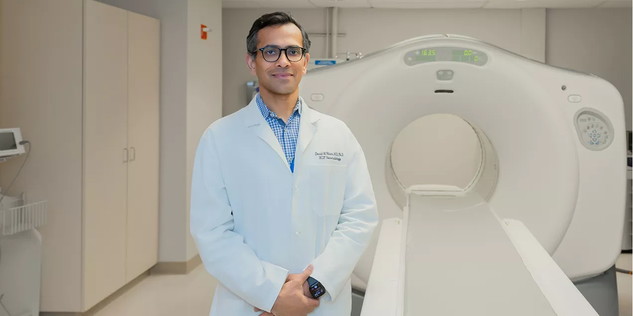 Radiologist in a white coat standing beside a CT scanner in a clinical imaging room, facing the camera.