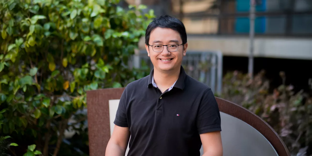 Portrait of a man with glasses smiling outdoors in a courtyard with greenery, wearing a black polo shirt
