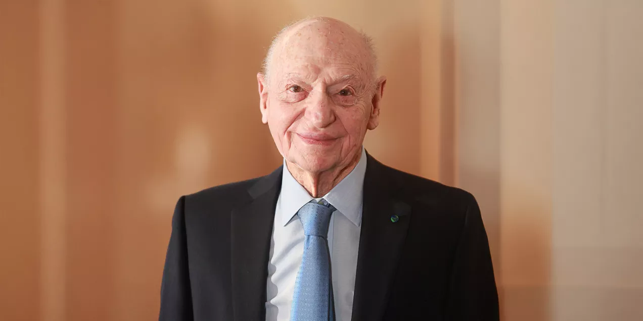 An elderly man with a gentle smile stands indoors wearing a dark suit, light blue shirt, and blue tie. He has a bald head with thin white hair at the sides and a warm, approachable expression. A softly lit sculpture is visible in the background, giving the setting a refined, professional feel.