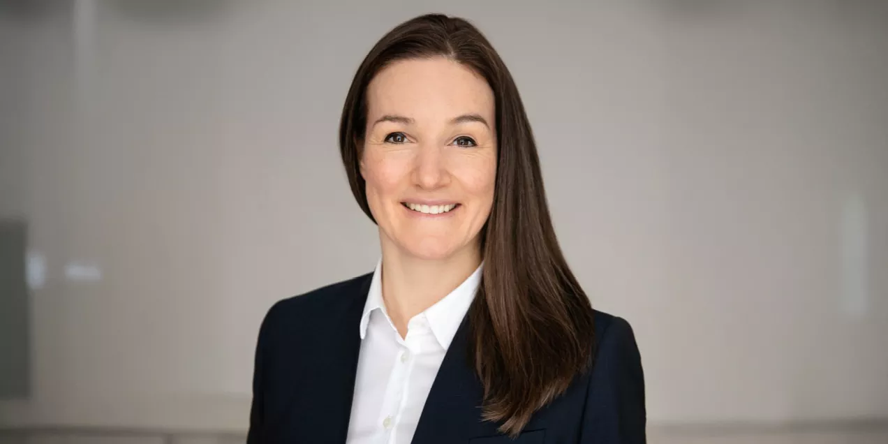 Professional headshot of a woman smiling at the camera. She has long, straight dark brown hair parted to one side and is wearing a dark navy blazer over a white collared shirt. The portrait is framed from the chest up against a soft, light gray background. The lighting is even and natural, creating a polished and approachable appearance suitable for a professional, academic, or corporate profile.
