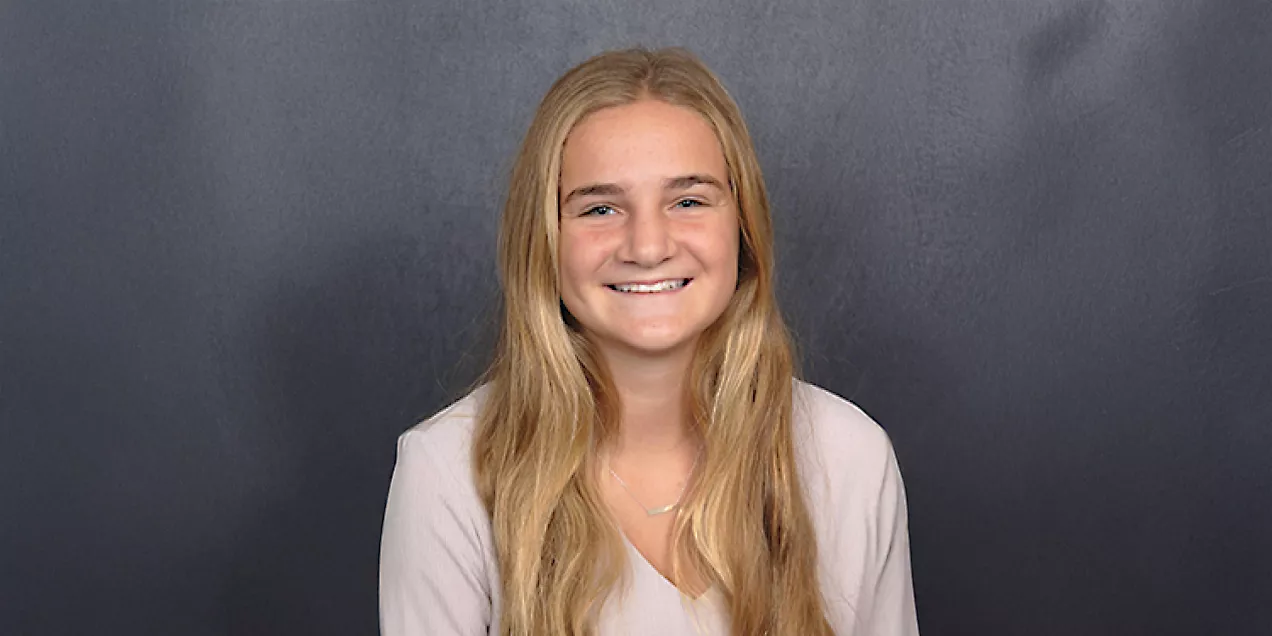 Portrait of Allison smiling at the camera against a dark gray background. She has long blonde hair and is wearing a light-colored blouse and a simple necklace.