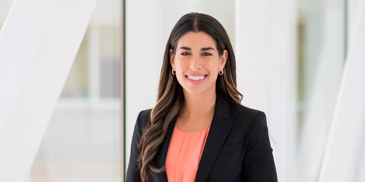 Professional portrait of a woman with long dark wavy hair wearing a black blazer over a coral blouse. She is smiling at the camera while standing in a bright, modern indoor space with large windows and softly blurred white architectural elements in the background.