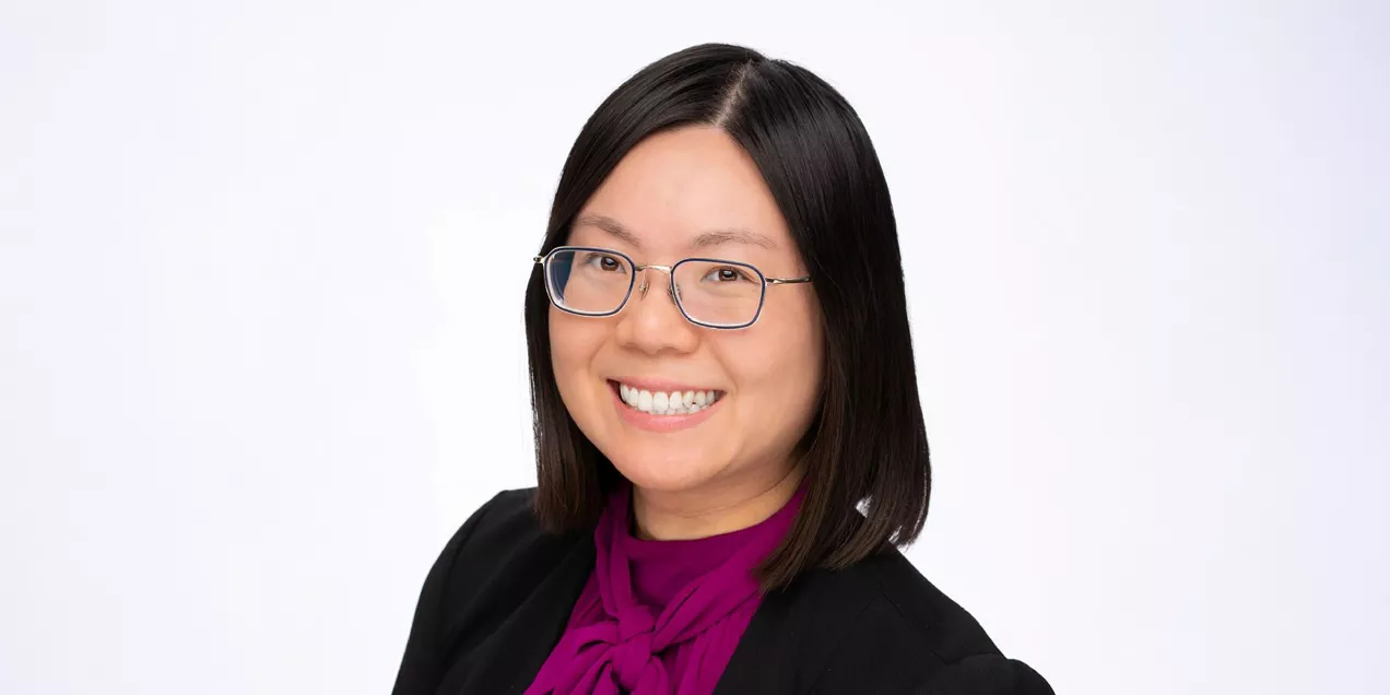 Professional headshot of a smiling woman with shoulder-length dark hair and glasses, facing the camera. She is wearing a black blazer over a magenta blouse with a tied neckline. The portrait is set against a light gray background, creating a clean and polished appearance suitable for a professional or faculty profile.