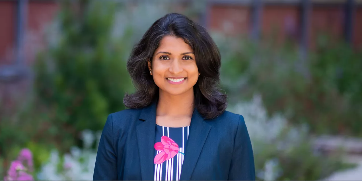 Professional outdoor portrait of a woman smiling at the camera. She is wearing a dark blazer over a striped top with a pink floral design. The background is softly blurred, featuring greenery, flowers, and a building, creating a natural campus setting.