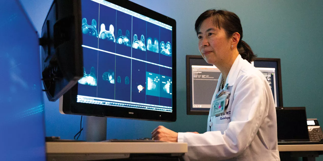 A radiologist reviews breast imaging scans displayed on a large medical monitor in a clinical reading room. Multiple breast MRI images are visible on the screen as she analyzes the results, highlighting the role of advanced imaging in breast cancer detection and diagnosis.