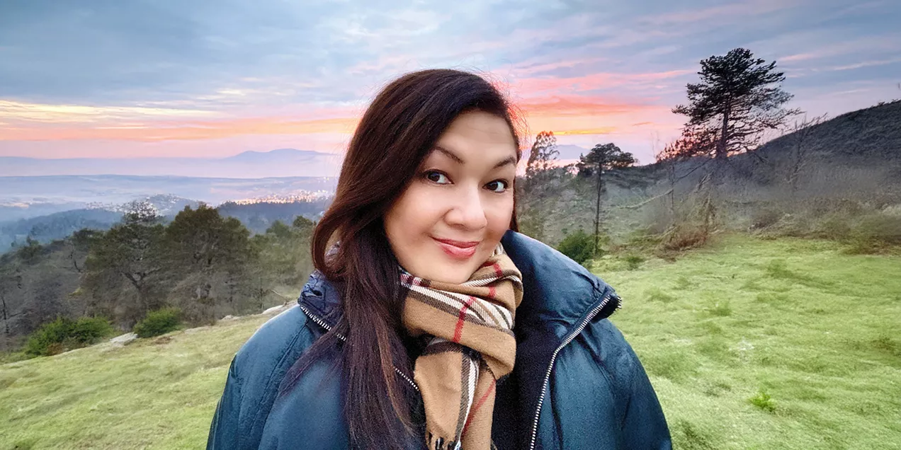 Portrait of a smiling person wearing a dark jacket and a tan plaid scarf, standing outdoors on a grassy hillside at sunrise. Trees, rolling hills, and a colorful sky create a scenic backdrop.