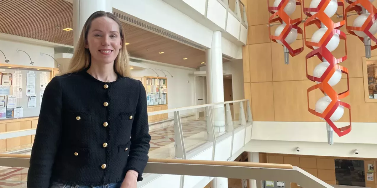 A woman stands in a bright, modern atrium, smiling at the camera while leaning against a railing. She wears a black textured jacket with gold buttons and blue jeans. The space features high ceilings, glass railings, warm wood paneling, and a large red-and-white hanging sculpture on the right side of the image. Bulletin boards and architectural details are visible in the background.
