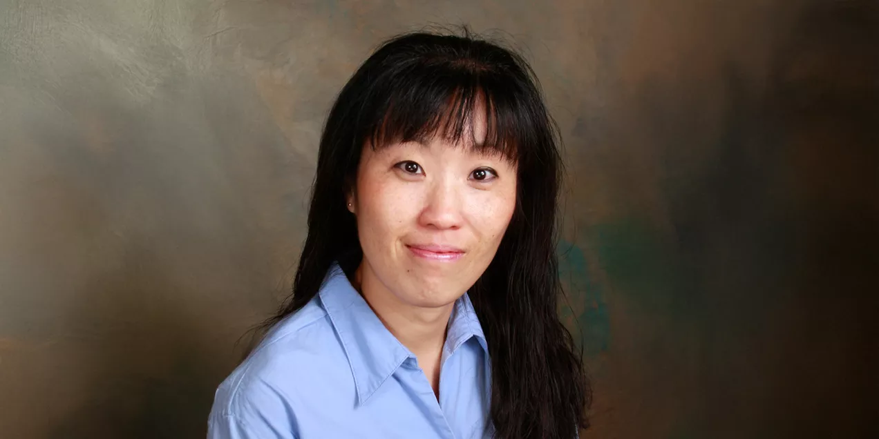 Professional portrait of a woman with long dark hair and bangs, wearing a light blue collared shirt. She is looking at the camera with a slight smile. The background is a softly blended studio backdrop in shades of brown and green, creating a warm and professional appearance.