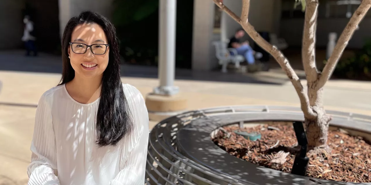 Portrait of a smiling woman with long dark hair and glasses seated outdoors in a sunlit courtyard. She is wearing a white blouse and looking toward the camera. Dappled shadows from nearby trees fall across her face and clothing. In the foreground is a circular bench surrounding a tree, while the background is softly blurred with building columns, walkways, and a person seated in the distance.