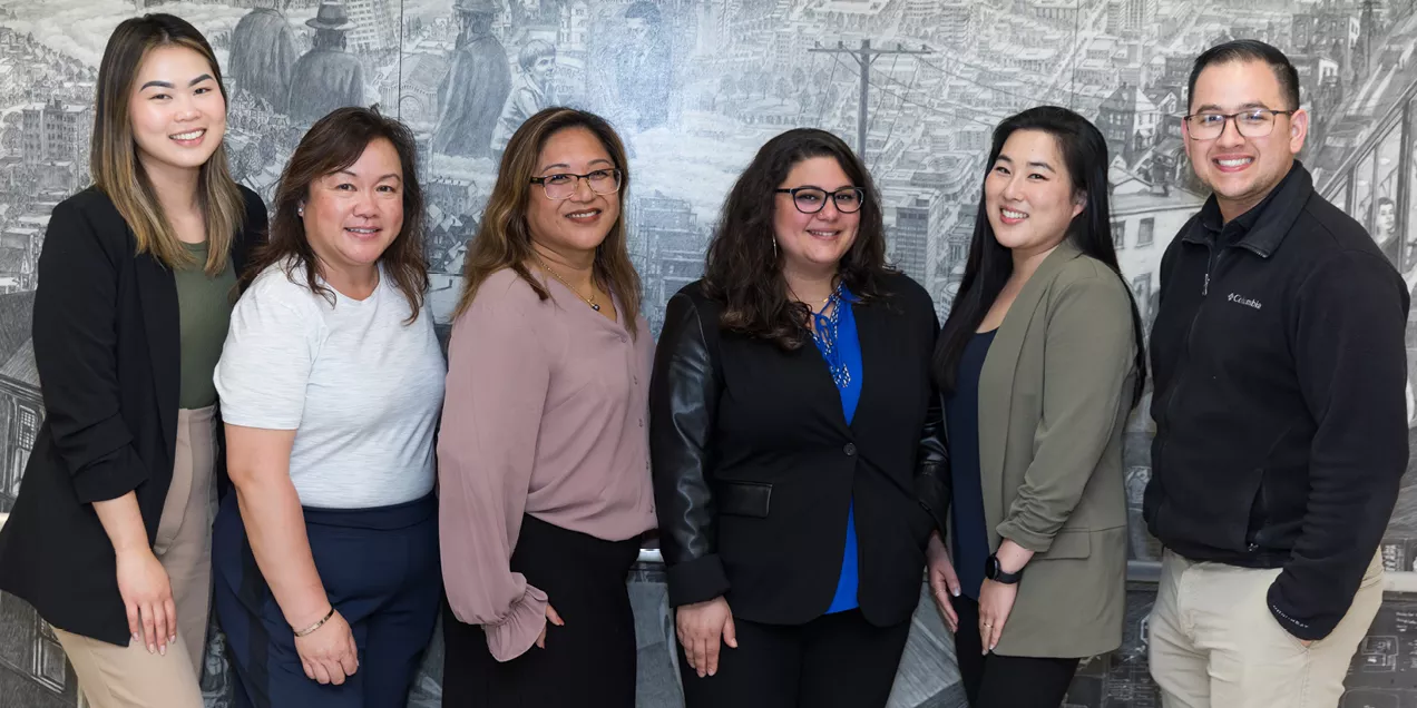 Group portrait of six smiling professionals standing side by side in front of a large black-and-white mural. The group includes five women and one man dressed in business or business-casual attire, with blazers, blouses, and collared shirts. They are facing the camera and posing for a professional photo, with the mural providing a detailed urban backdrop.