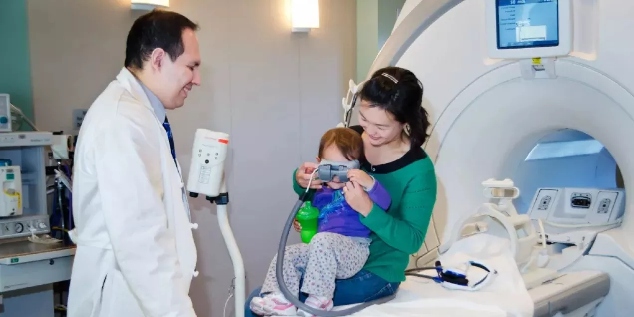 A healthcare provider stands beside an MRI scanner while a parent holds a young child sitting on the MRI table. The child is wearing video goggles, likely to stay calm and watch content during the scan, while the parent smiles and assists.