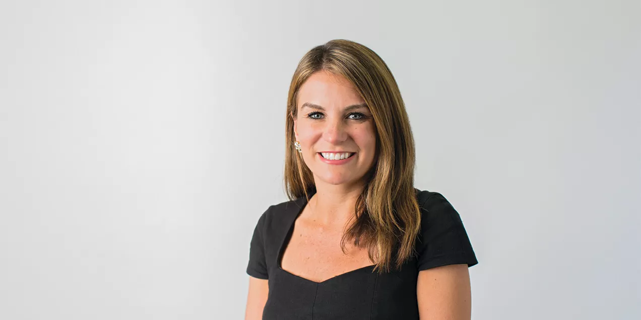 Smiling woman with long light brown hair wearing a black short-sleeved dress, posed against a light gray background. She is facing the camera with a friendly expression and wearing small hoop earrings. Professional studio headshot.