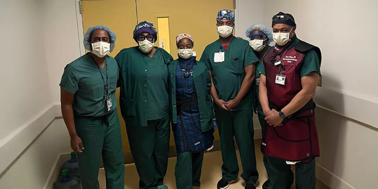 Six healthcare professionals wearing surgical scrubs, masks, and protective caps stand together in a hospital hallway outside an operating room. One team member is wearing a lead apron, and the group is facing the camera for a team photo.
