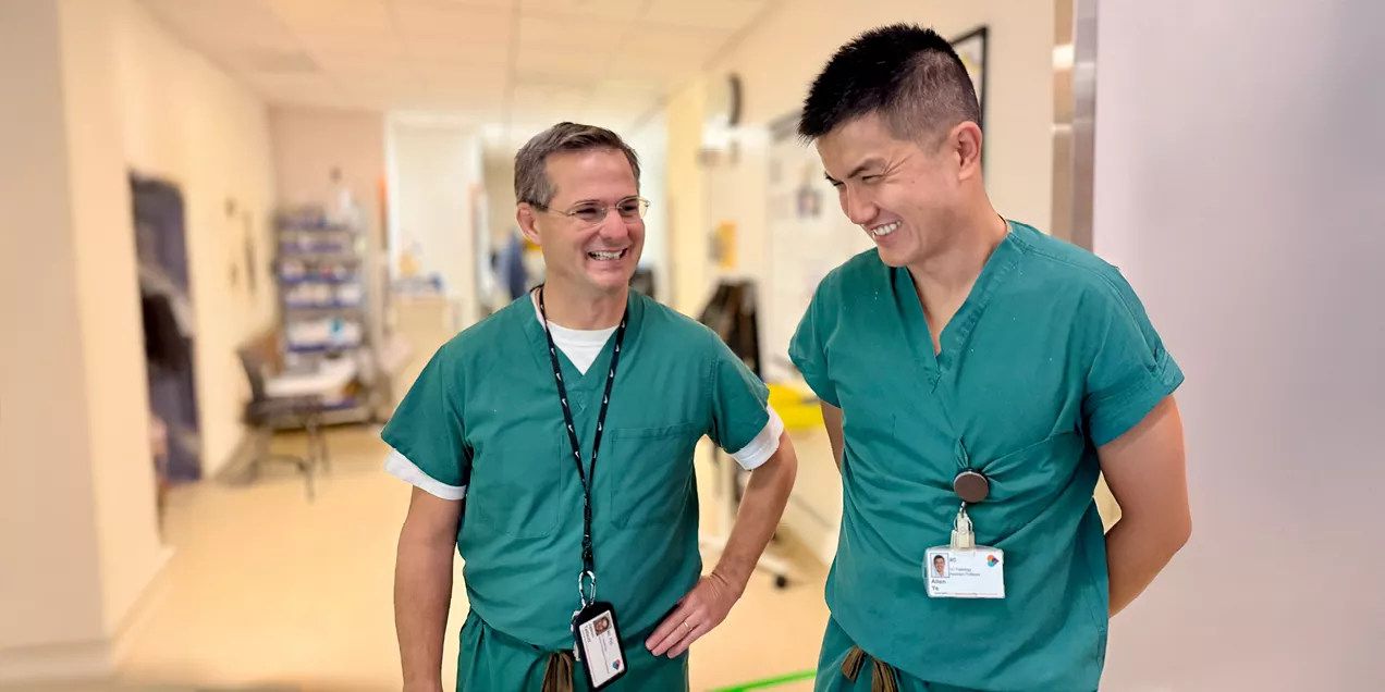 Jason Talbott and Allen Ye, both wearing green surgical scrubs, smile and talk while standing in a hospital hallway. The candid moment captures collegiality and teamwork in a clinical care setting.