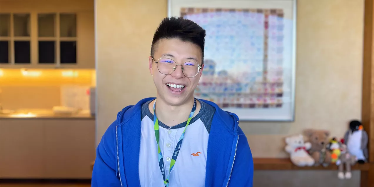 Smiling person wearing round glasses, a blue zip-up hoodie, and a conference lanyard, standing indoors in front of framed artwork. Plush toys are displayed on a shelf in the softly blurred background.