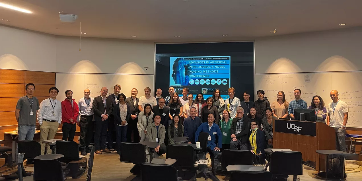 Group photo of conference attendees gathered in a UCSF lecture hall. Participants stand and sit in front of a presentation screen displaying a conference on artificial intelligence and novel imaging methods. The room includes classroom seating, whiteboards with notes, and a UCSF podium, capturing researchers, faculty, students, and guests at the conclusion of the event.