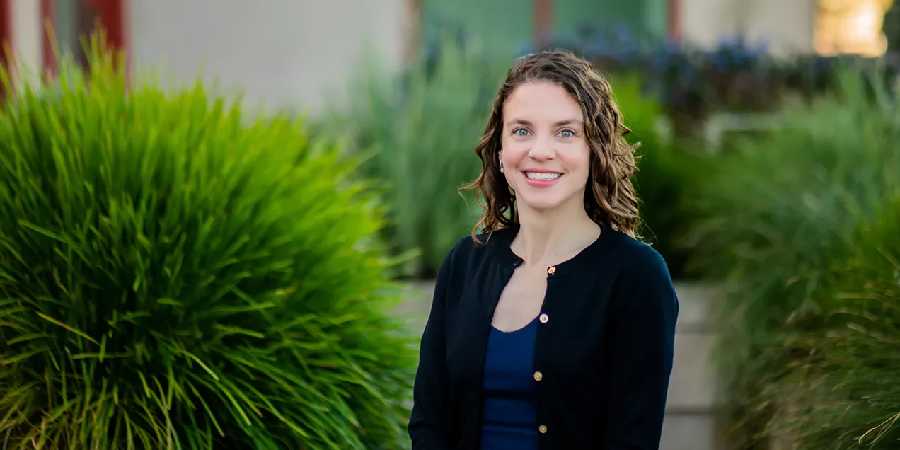 Portrait of Kimberly Kallianos, MD, smiling outdoors, seated in front of softly blurred green landscaping.