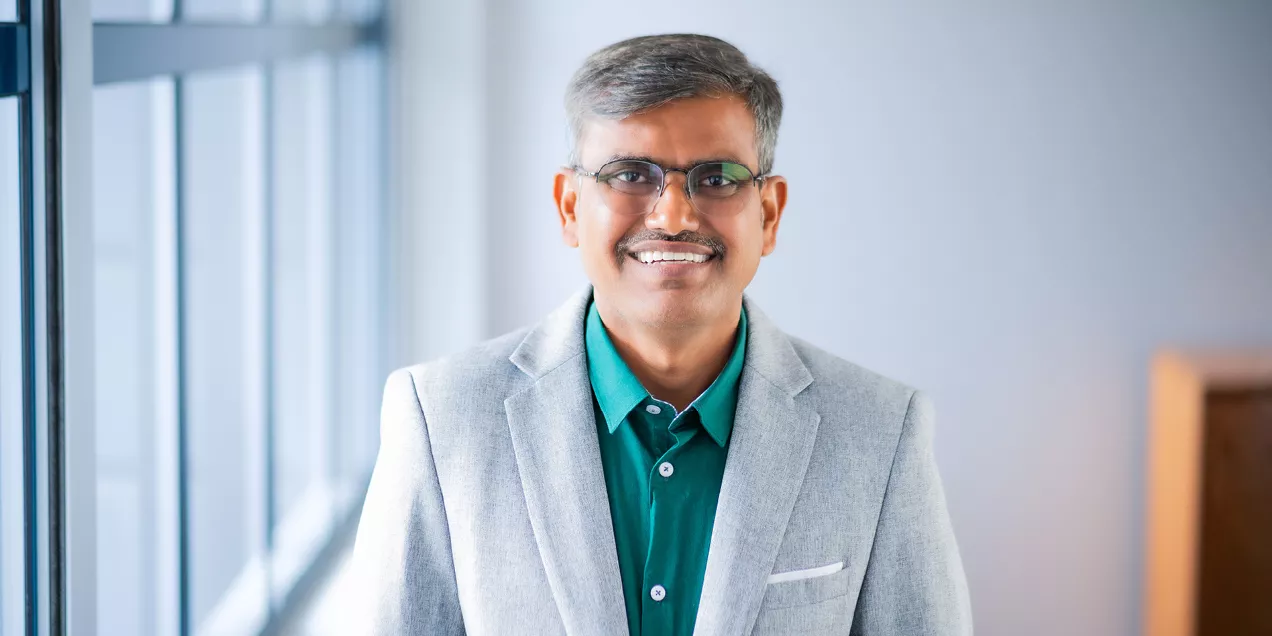Professional portrait of Kondapa Naidu Bobba smiling while standing indoors near a window. He is wearing glasses, a light gray blazer, and a teal shirt, with soft natural light and a blurred background creating a welcoming and professional appearance.