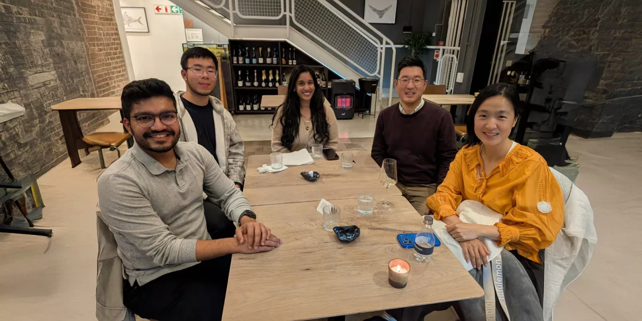 Five people sit around a wooden table in a modern restaurant or café, smiling toward the camera. The group appears to be enjoying a casual social gathering, with glasses, drinks, and a lit candle on the table. The space features exposed brick and stone walls, a staircase in the background, and warm indoor lighting that creates a welcoming atmosphere.