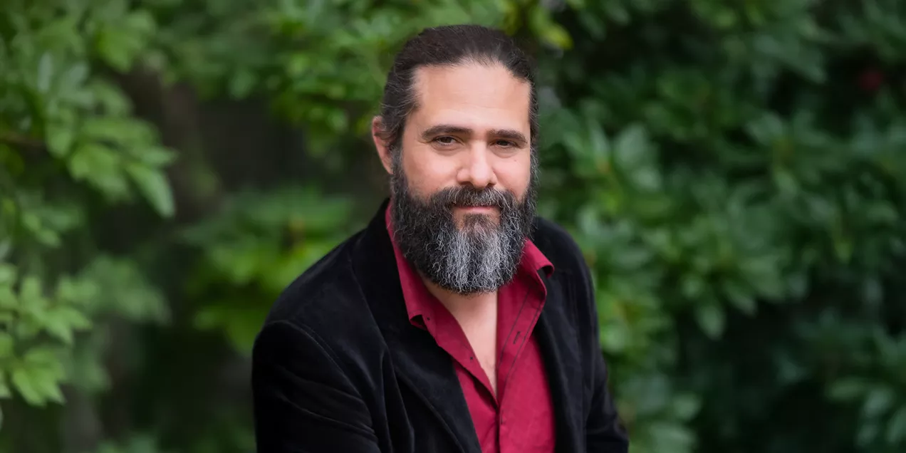 Portrait of a bearded man with long hair pulled back, wearing a red button-down shirt and black blazer. He is seated outdoors and looking at the camera, with green foliage softly blurred in the background.